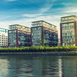 3D rendering of 4 rectangular buildings on a sunny day with a tree-lined walkway dividing it from a river